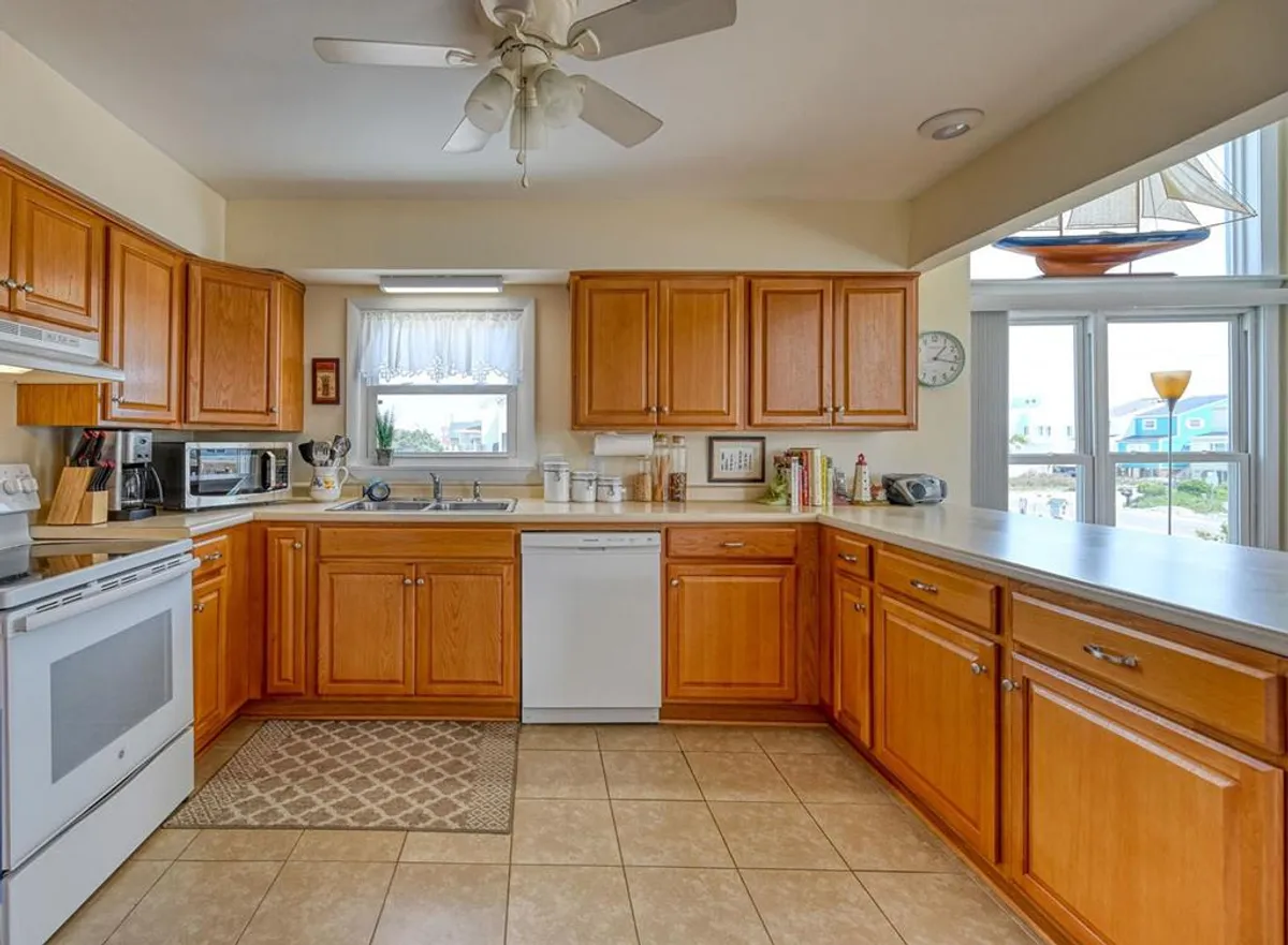 A bright, inviting kitchen is shown, with wooden cabinets, kitchen appliances, and a window letting in natural light. Decorative touches like a sailboat model create a coastal vibe, and the space is clearly designed for cooking and gathering.