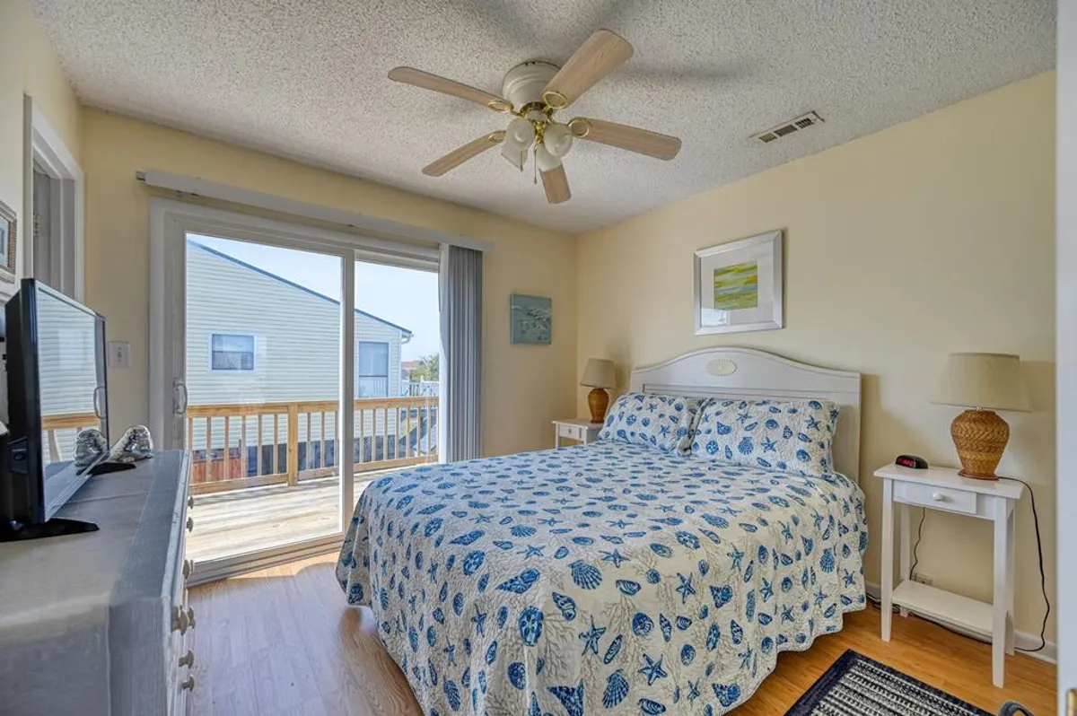 This image features a bedroom with a bed dressed in blue and white patterned bedding, a sliding glass door leading to a private balcony, and a ceiling fan overhead. Soft natural light streams into the room, creating a serene and inviting atmosphere ideal for rest and rejuvenation.
