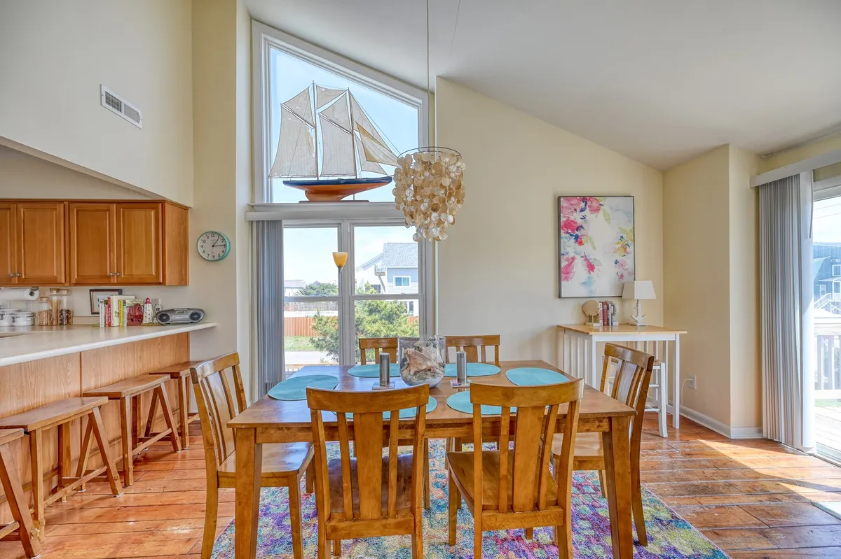 A warm dining area with a large table set for six, a chandelier overhead, and a window decorated with a sailboat figure. Colorful artwork adds a cheerful touch, while the window frames provide a bright, airy atmosphere and glimpses of the outside.