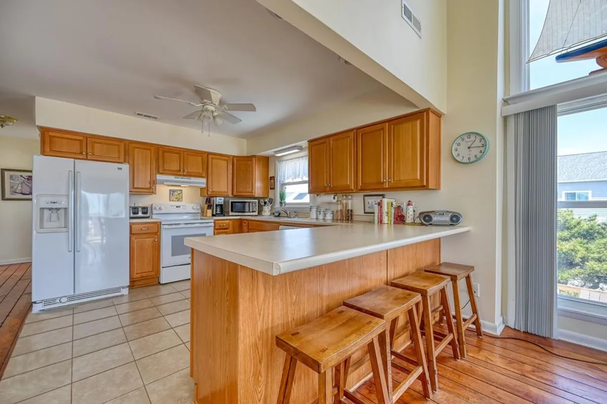 A bright, welcoming kitchen with a central island featuring wooden bar stools, white appliances, and natural wood cabinetry. The scene shows a functional, fully equipped kitchen space that's both practical and inviting for meal preparation or casual dining.