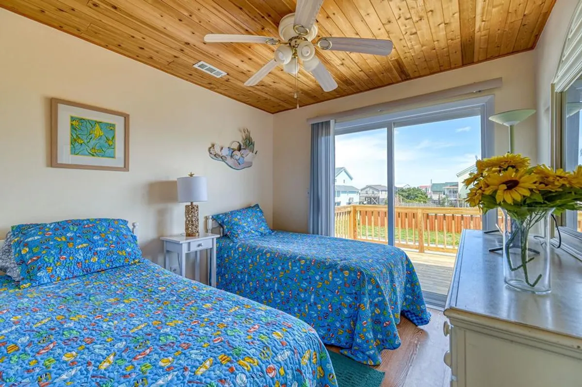 A guest bedroom featuring two twin beds with blue floral bedding, a white dresser, a sliding glass door to the deck, and a bedside lamp. The space is bright and cheery, emphasizing both comfort and a fresh coastal aesthetic.