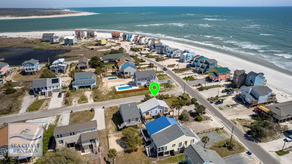 This image features a beachfront with colorful houses lining the shore and a swimming pool in the foreground. The ocean is visible beyond, conveying the proximity of the property to both the beach and water-related amenities. The setting radiates a relaxed vibe, ideal for family gatherings, sunbathing, swimming, or beachcombing.