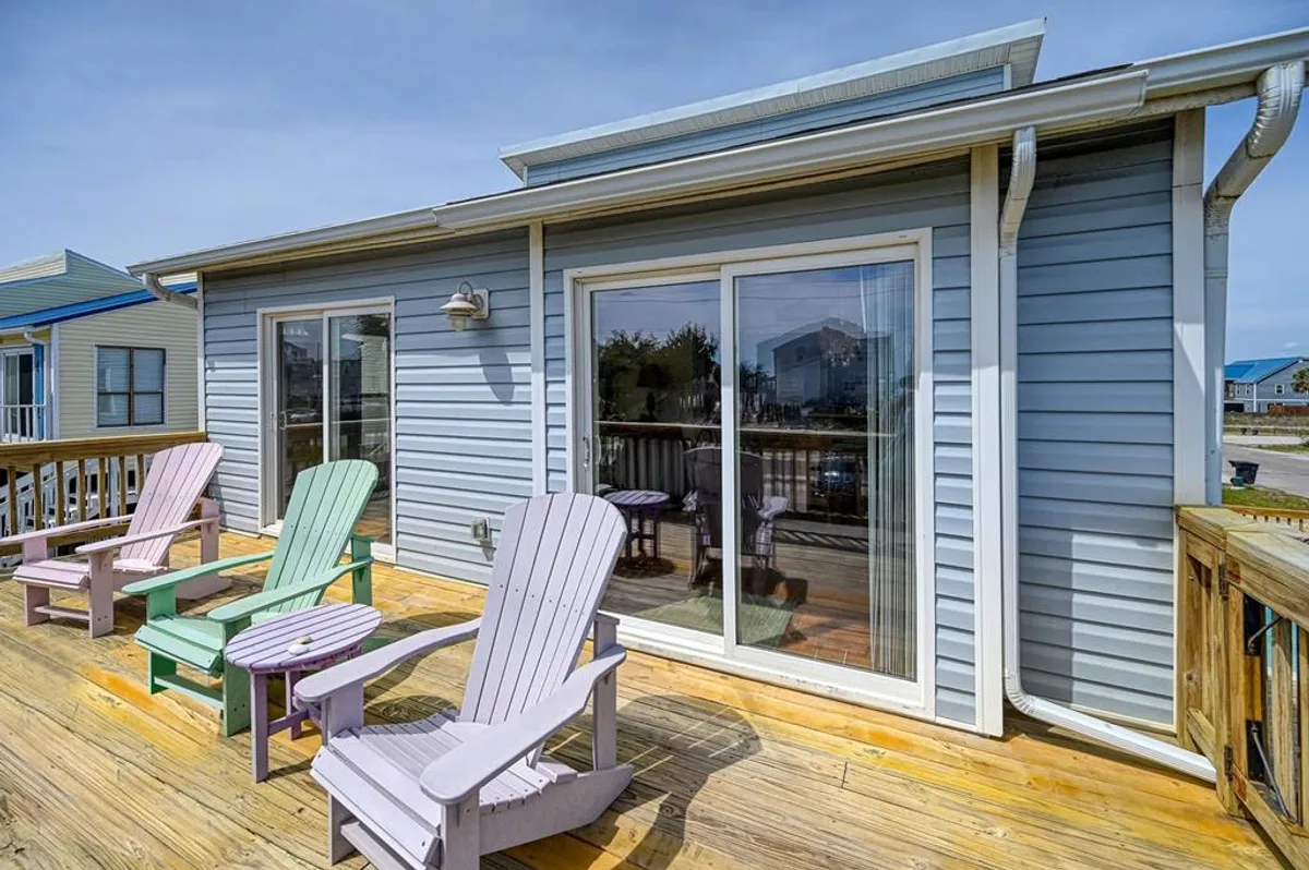 This image captures a tranquil exterior deck with colorful Adirondack chairs, glass sliding doors, and a wooden deck bathed in natural light. The inviting scene looks out toward the ocean, suggesting an ideal spot for morning coffee or sunset relaxation, with easy access from the primary living area.