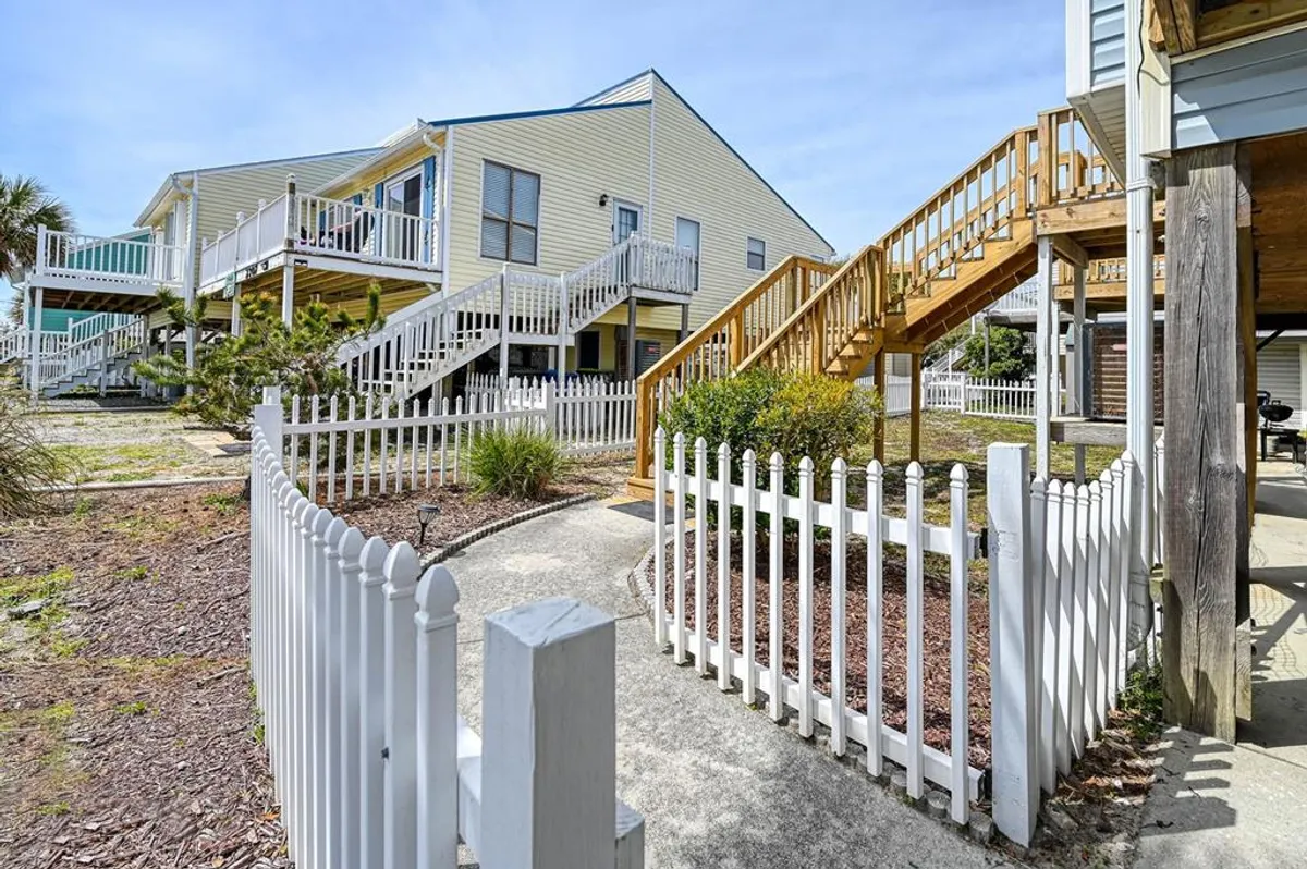 An exterior image featuring a wooden staircase leading up to the main level of the home, surrounded by white picket fences and landscaped pathways. The stairs are prominent, making clear the need to climb to the main living space.