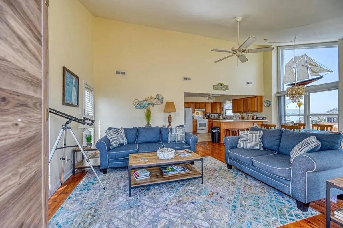 This interior image captures comfortable blue sofas centered in a sunlit living area, a wood coffee table, and a decorative sailboat hanging from the vaulted ceiling. A telescope sits near the window, emphasizing the ocean views beyond. The mood is casual and inviting, perfect for relaxation or entertaining guests.
