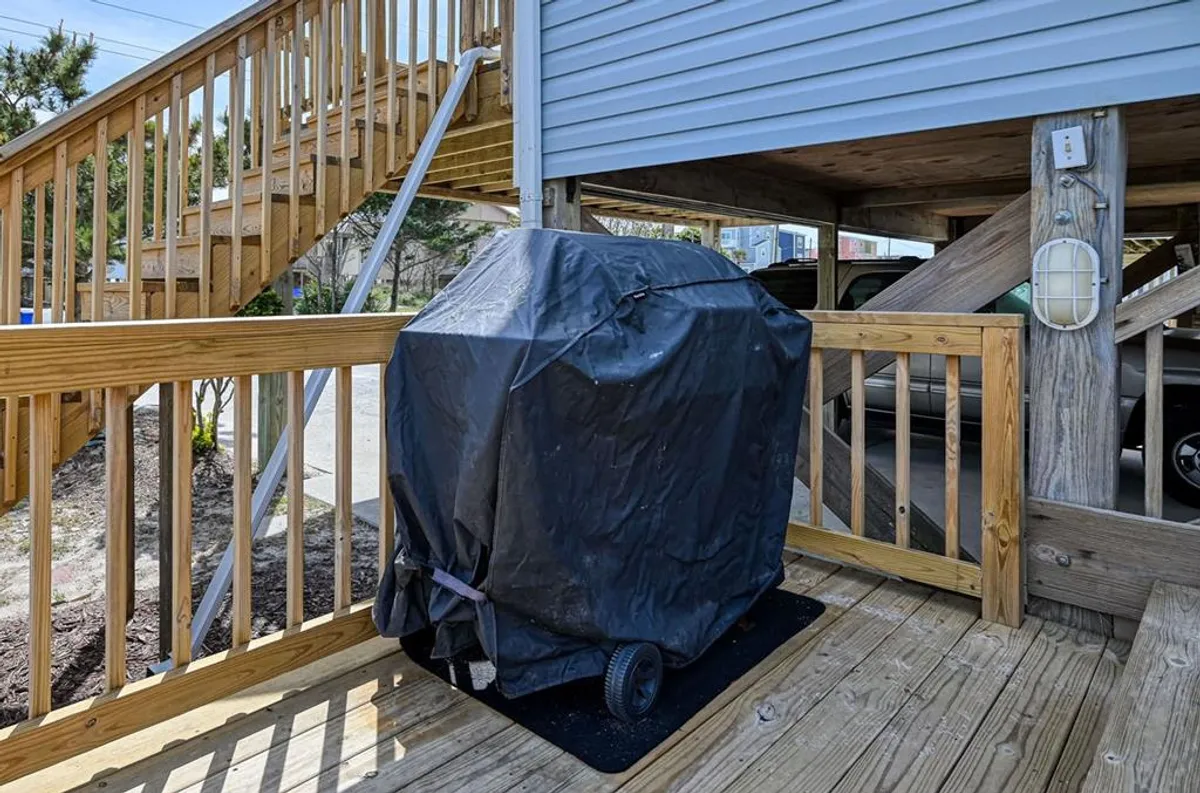 The photo shows an exterior deck with a grill covered by a black tarp, wooden deck railing, and stairs leading up, indicating an elevated living area. The setting suggests casual, communal activities such as barbecuing and outdoor meals with family or friends.