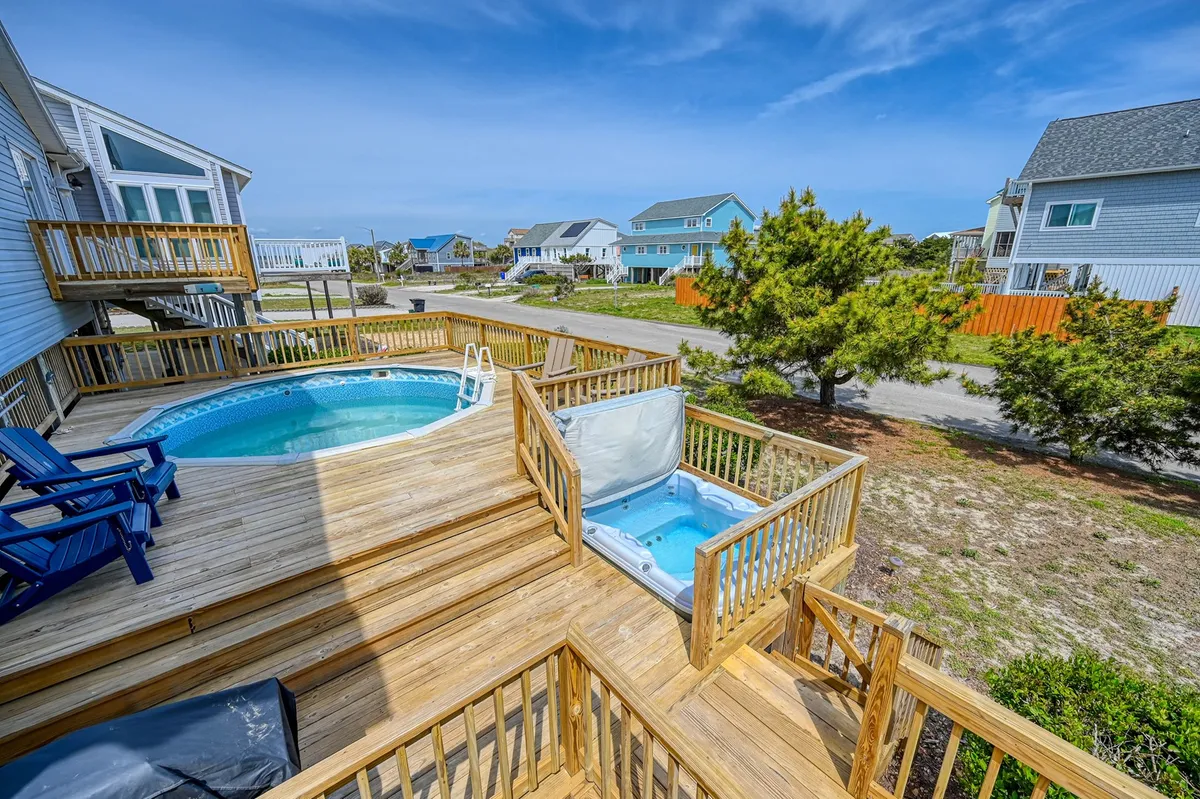 The image showcases the inviting exterior of a beach house, with a sparkling swimming pool and bubbling hot tub bordered by a spacious wooden deck. Blue chairs are arranged for lounging and socializing, all set under the serene North Carolina sky. The scene radiates relaxation and leisure, with the pool and hot tub as the central highlights, suggesting both daytime fun and evening unwinding.