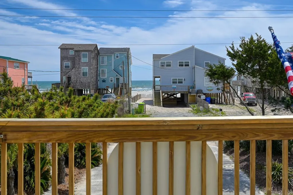 A scenic view of beachfront houses with tall palm trees and the ocean stretching beyond. The image conveys the experience of being surrounded by the relaxing sights and sounds of the coast, perfect for beachcombing, fishing, sunbathing, or taking a sunset stroll.