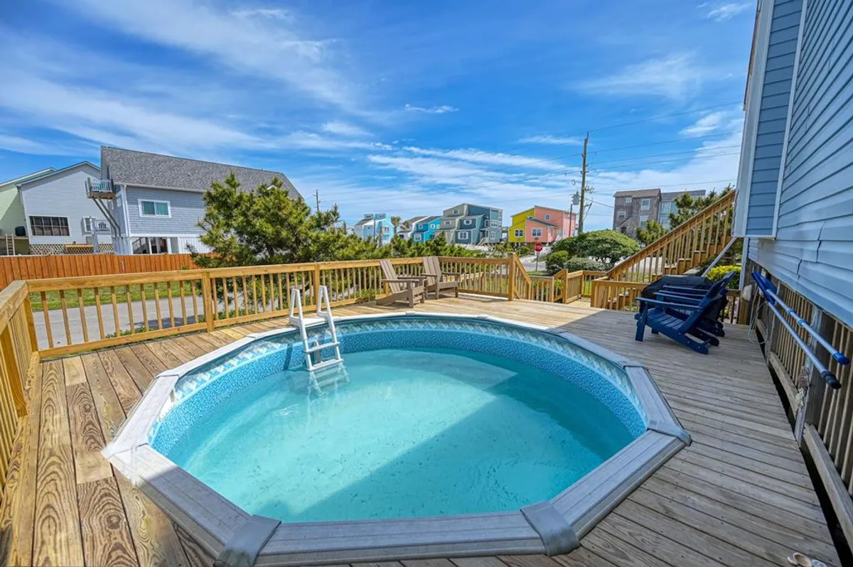 A bright swimming pool area surrounded by a spacious deck and lounge chairs, with colorful houses in the background. The scene radiates with energy and leisure, ideal for swimming, sunbathing, and poolside fun.
